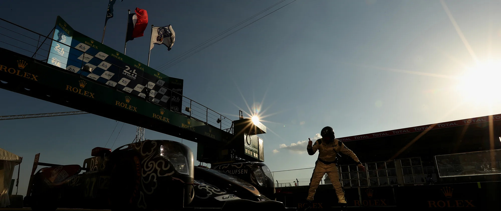 A race car driver giving a thumbs up beside a race car