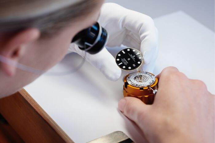 A watchmaker in white gloves carefully assembles a luxury Rolex watch, holding its black dial and golden case.